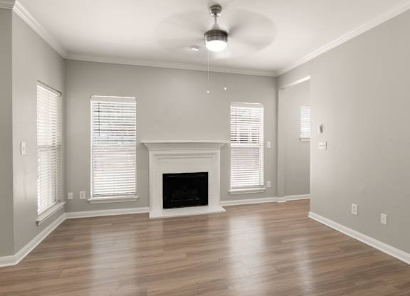 Open living room with wood-style flooring, white fireplace mantel, ceiling fan, and tall windows.