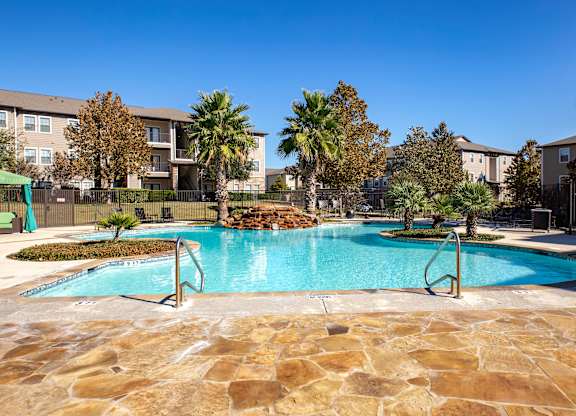 Curved community pool with stone deck, palm trees, and nearby lounge seating.