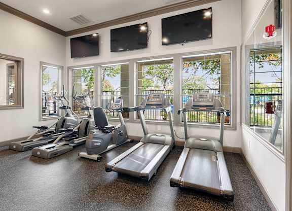 Fitness center with treadmills, bikes, ellipticals, tall windows, and wall-mounted televisions.