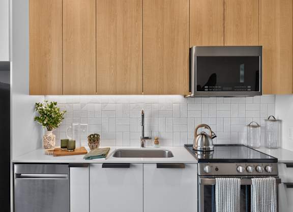 A modern kitchen with a stainless steel refrigerator, a microwave above the stove, and a sink with a kettle on it at The Indie in Allston.