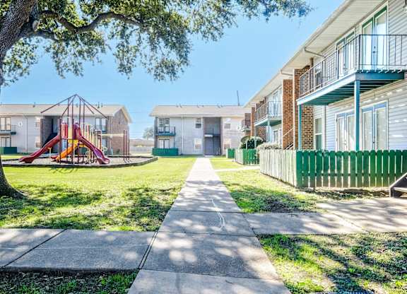 A playground with a slide is in the middle of a grassy area between two apartment buildings.