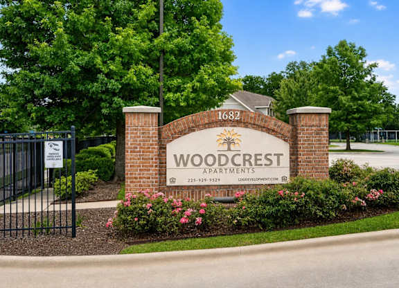 The image shows a sign for Woodcrest Apartments with a black fence and flowers in the foreground.