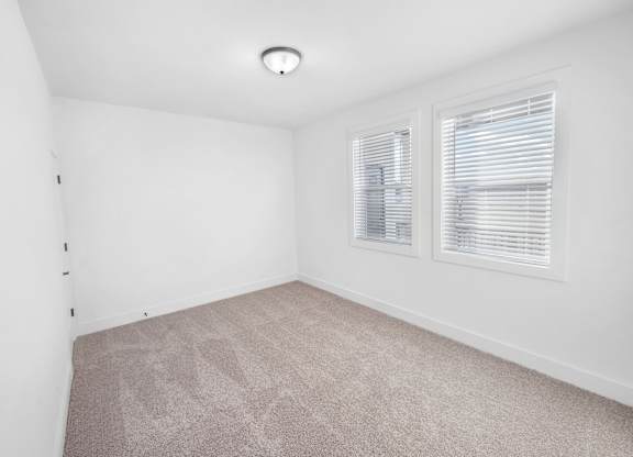 A room with a carpeted floor and two windows with blinds.