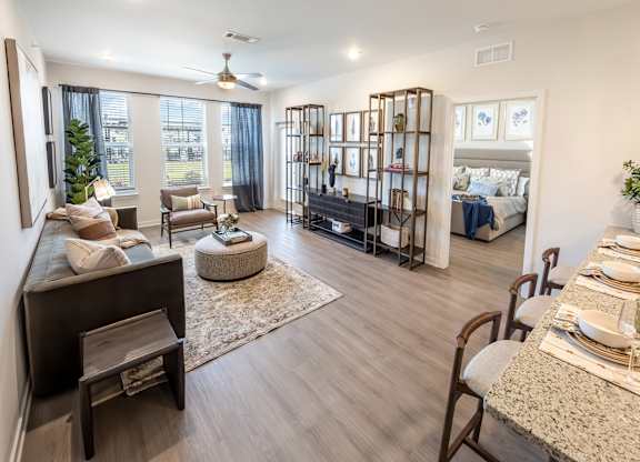 A well-lit, spacious living room with a large window, a sofa set, and a dining area with a table set for two.