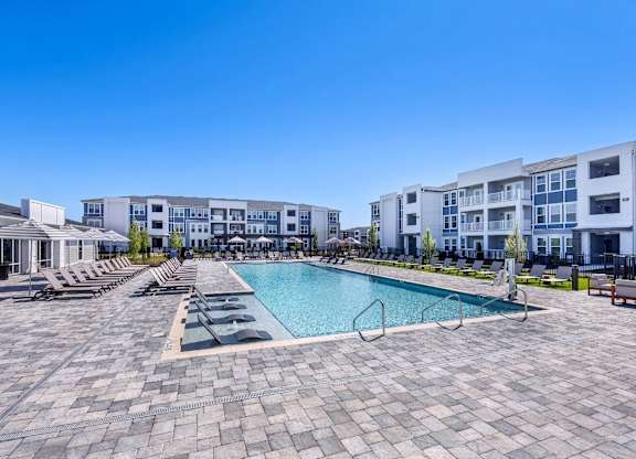 A large swimming pool surrounded by lounge chairs and buildings in the background.