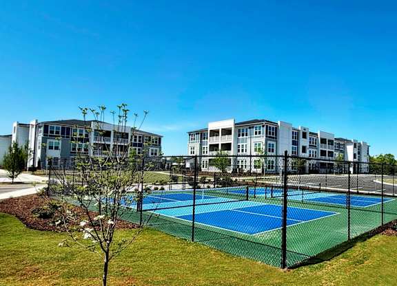 A tennis court is surrounded by a fence and a grassy area in front of a building.