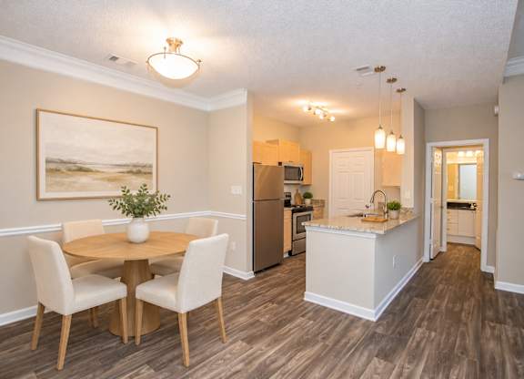 A dining room with a table and chairs and a kitchen in the background.