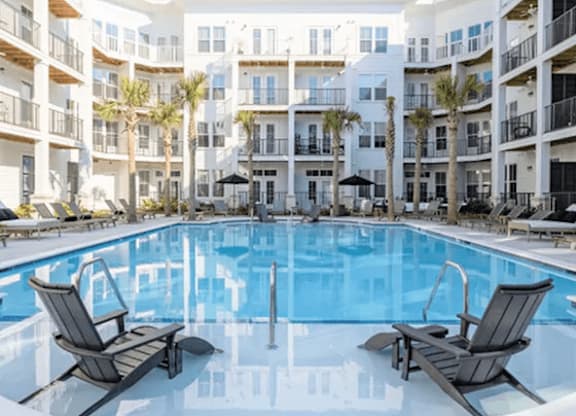 A swimming pool with two lounge chairs in front of a white apartment building.