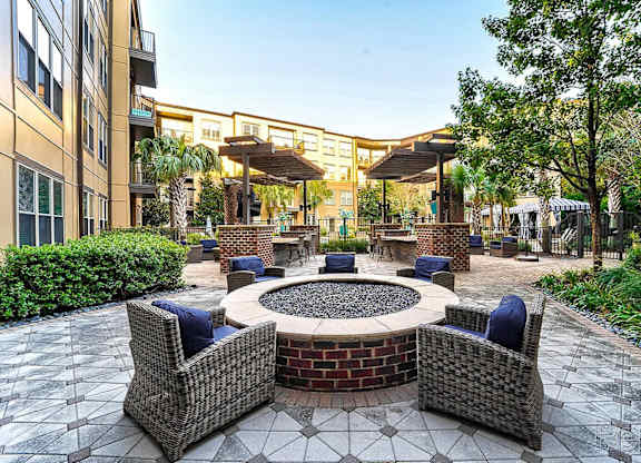 A patio with a fire pit surrounded by chairs and a building in the background.