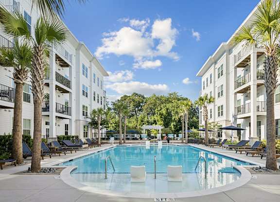 A swimming pool surrounded by palm trees and lounge chairs in front of a white apartment building.