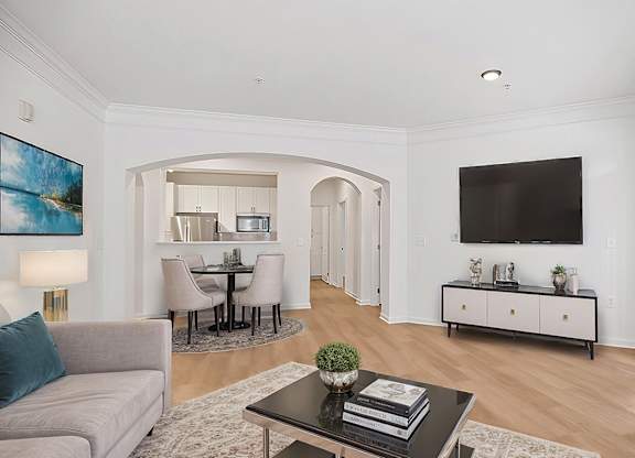 A living room with a grey couch, a coffee table, a dining table, and a television mounted on the wall.