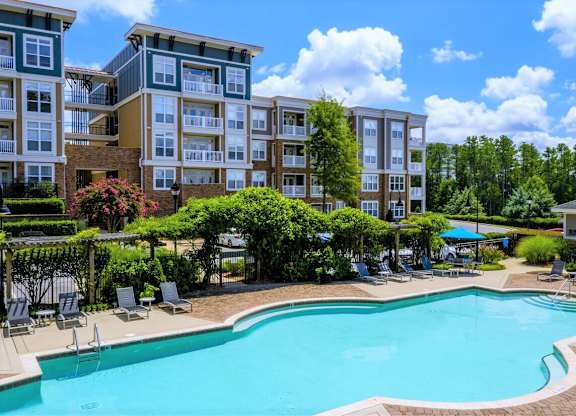 A swimming pool surrounded by lounge chairs and trees in front of apartment buildings.
