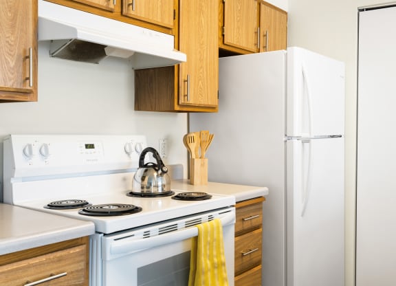 The corner of the kitchen with a white oven, stove top, hood range, and full-size fridge. Staged with a tea kettle, wooden spoons, and a hand towel.at Clearwater, Post Falls