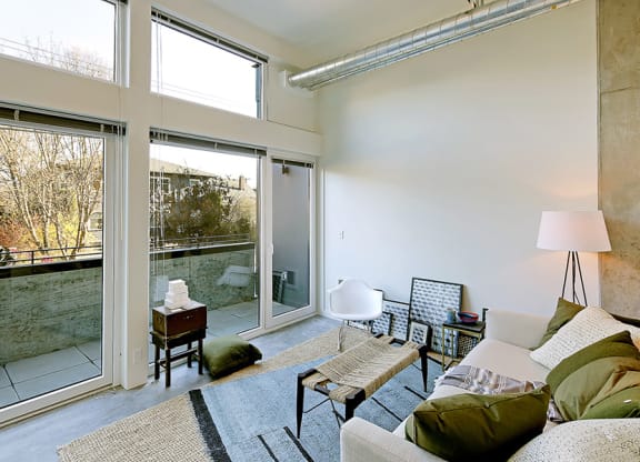 Living room with concrete flooring, floor to ceiling windows and a private outdoor patio. Inside is staged with a coffee table, rug and chair.at 19th & Mercer, Washington