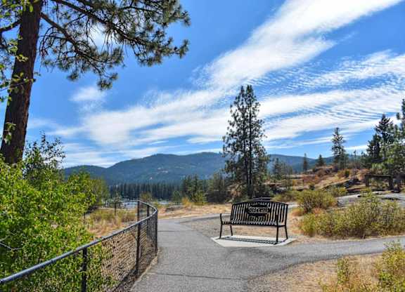 A park bench sits on a paved path with a metal fence and trees in the background.