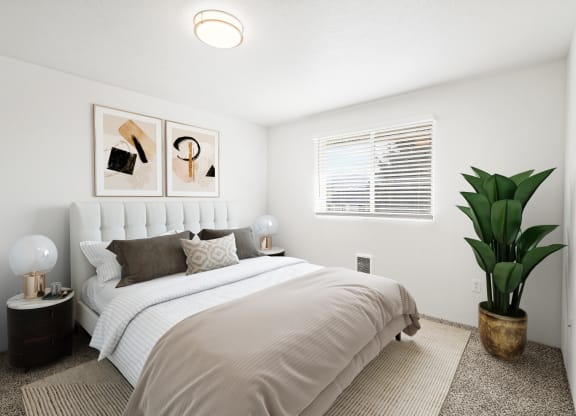 a large bedroom with white walls, taupe carpet, and a window on the back wall. Staged with queen-size bed, 2 side tables, art and a plant.at North Pointe, Post Falls