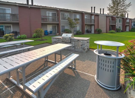BBQ and picnic area with two picnic tables, a stainless steel BBQ, and a large green lawn and landscaping behind. at Pointe East, Fife, WA, 98424