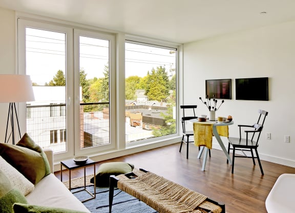 Living room with floor to ceiling windows with city and tree views. Staged with a couch, coffee table, rug and chair.at 19th & Mercer, Seattle Washington