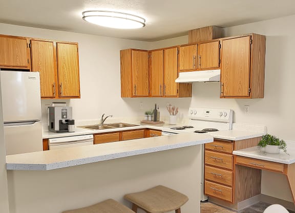Kitchen with bar counter and stools, wood cabinets, white appliances, a coffee station, and house plants.at Clearwater, Post Falls, 83854