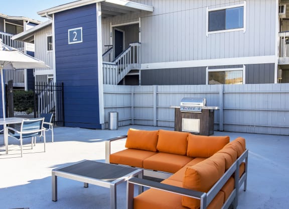 Outdoor lounge with umbrella-covered tables, gas BBQ, and an oversized orange patio couch and coffee table.at The Lakes Apartments, Moses Lake, WA