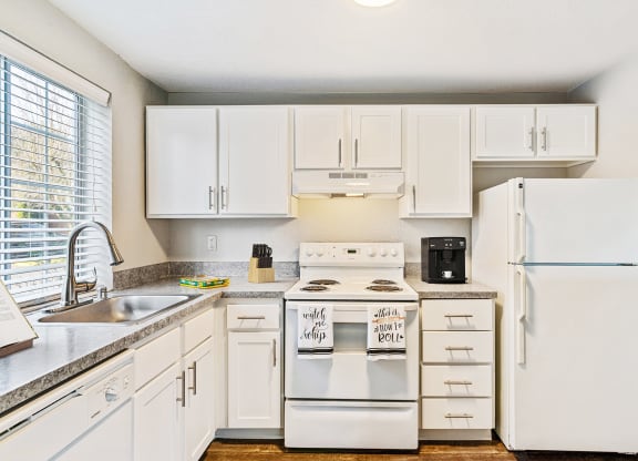 L-shaped kitchen with white cabinets, a large window above the sink, and a white dishwasher, oven, stovetop, full-size fridge.at Capitol Crossing, Olympia, WA