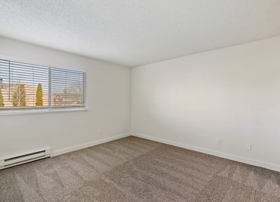 A spacious bedroom with white walls, sunny windows, a heater below the window, and taupe carpets. at Pointe East, Washington, 98424