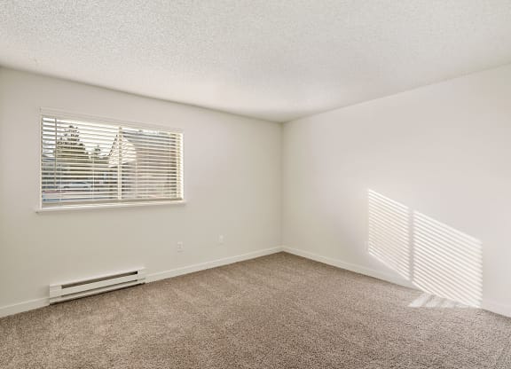 A large bedroom with white walls, taupe carpets, and a sunny window with white blinds. at Pointe East, Fife, Washington