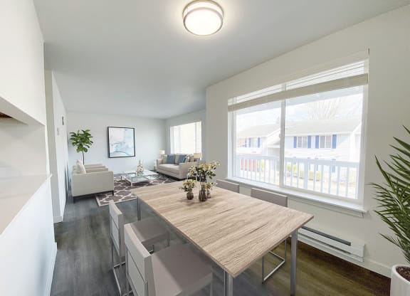 A dining room with white walls and a large sunny window that looks into the living room. Light-filled apartment staged with dining set and living room furniture.at The Trail, Snohomish, WA