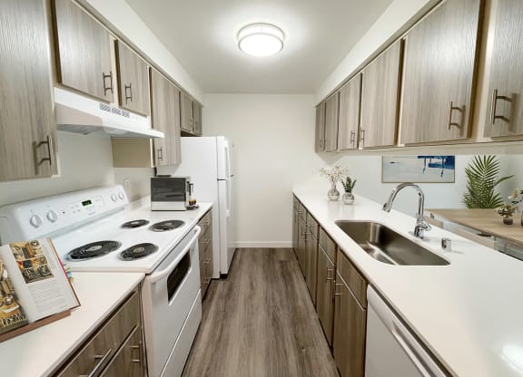 A galley-style kitchen with wood-like flooring, top and bottom cabinet storage, and white appliances. An oven and range, full-size fridge, dishwasher and sink.at The Trail, Snohomish, WA 98290