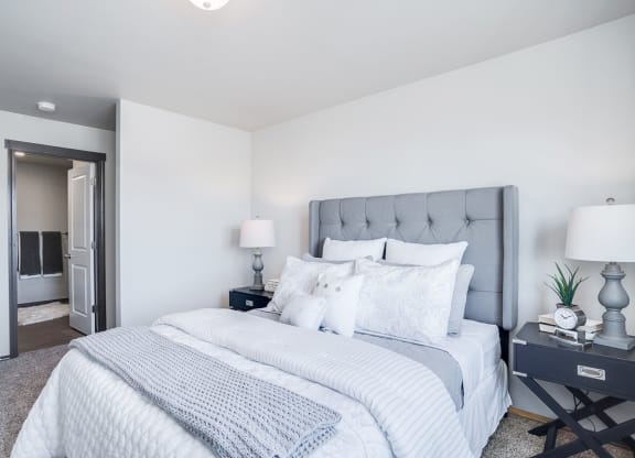 Large carpeted bedroom with full ensuite bathroom on the left. Staged with a queen bed, side tables, and lamps.at North Pointe, Idaho