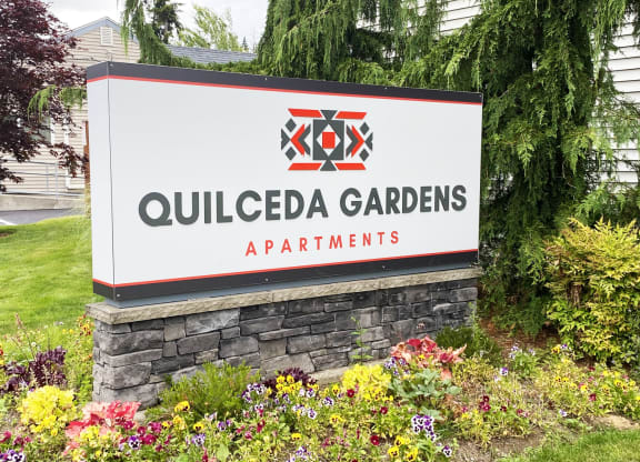 Community entrance with red and gray 'Quilceda Gardens' sign surrounded by flowers, grass, and trees.at Quilceda Gardens, Marysville