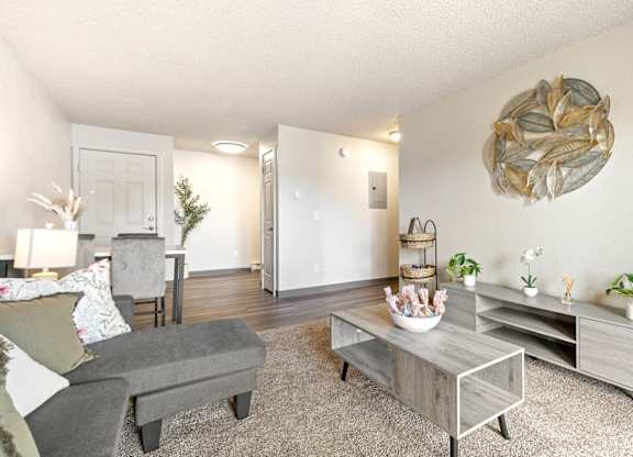 A living room with a grey couch, a coffee table, and a large sculpture on the wall A row of apartment buildings with green lawns in front A bedroom with a bed, nightstand, and window at Wildflower, Kennewick, WA 99336..