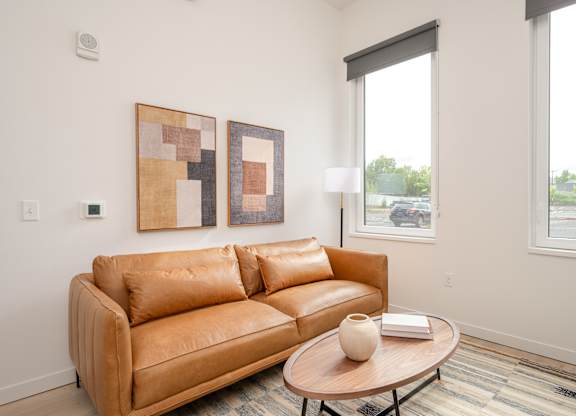 A tan couch with a white vase on a coffee table in front of a window.