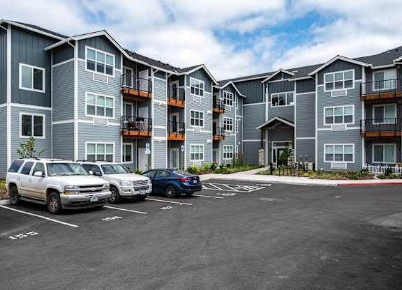A parking lot in front of a grey apartment building with two cars parked.