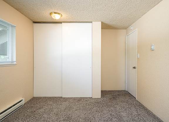 A room with a carpeted floor, a window with blinds, a white door, and a light fixture on the ceiling.