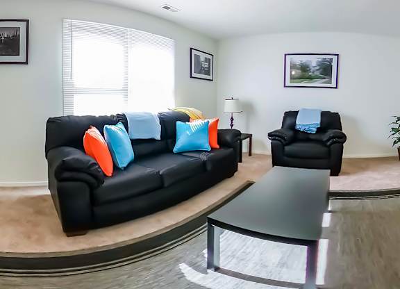 A black couch with orange and blue pillows sits in a living room with a black armchair and a coffee table.