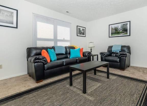 A black leather couch with orange and blue pillows sits in a living room with a coffee table in front of it.