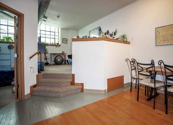 A room with a wooden floor and a white wall with a shelf and chairs.
