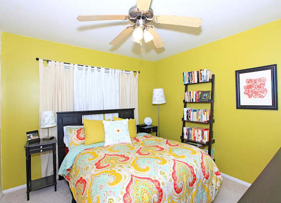 A bedroom with a bed, a ceiling fan, a nightstand, a lamp, a bookshelf, and a picture on the wall.