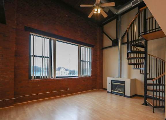 A room with a wood floor, a brick wall, a ceiling fan, and a staircase.