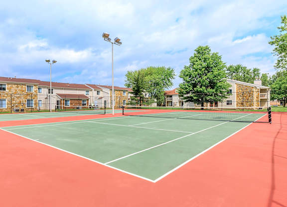 A tennis court with a red surface and white lines is surrounded by apartment buildings.