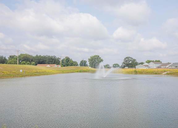 A fountain in the middle of a lake with houses and trees in the background.