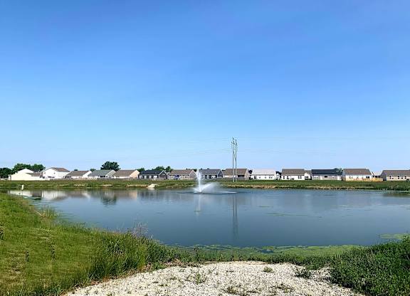 A serene lake with a fountain in the middle, surrounded by houses and a clear blue sky.