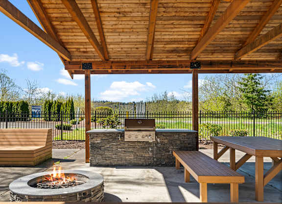 A wooden picnic table is under a wooden pergola with a fire pit in the middle.