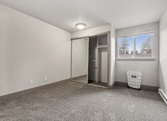 A room with a grey carpet and a white wall with a window and a trash bin.