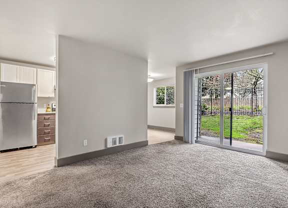 A spacious, empty room with a grey carpet and a sliding glass door leading to a green outdoor area.