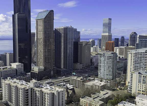 A cityscape with a mix of modern and older buildings, a body of water in the background, and a partly cloudy sky.
