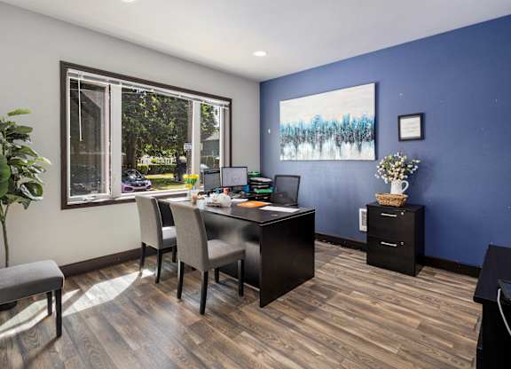 A room with a desk and chairs with a window and a painting on the wall.