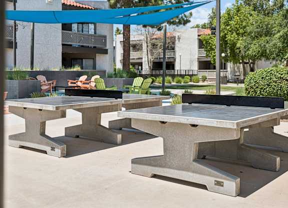 A concrete table with a blue canopy is in the foreground of a sunny outdoor area.
