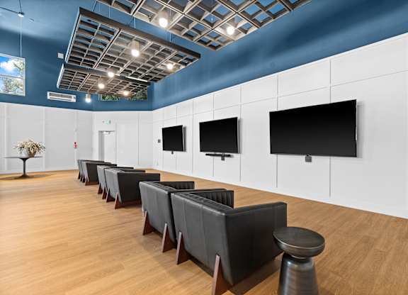 A room with a long row of chairs and three TVs on the wall.
